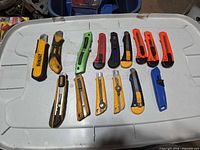 15+ assorted snap-off utility knives laid out on plastic bin lid