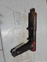 Right-angle pneumatic needle scaler showing metal housing, bundle of steel needles, ribbed plastic grip, rust