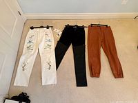 Three pairs of pants laid flat: white embroidered jeans, black panelled pants with tags, brown jogger pants