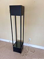 Full view of black metal and glass floor lamp showing column frame, glass shelf, power cord