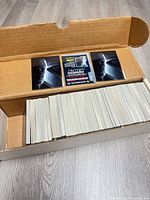 Open cardboard box showing large stack of trading cards with two cards displayed on top