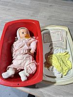 Baby doll lying in red plastic bin, lid alongside showing paperwork and yellow garment