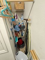 Narrow laundry closet showing shelves with iron, bleach, cleaners, hangers, ironing board, buckets and hamper