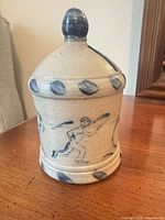 Front view of stoneware coin bank showing lid, knob finial and hand-painted figure