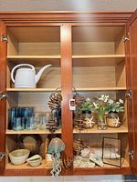 Cabinet showing watering can, blue glasses, tumblers, decorative jars with flowers, wind chimes, jellyfish ornament, bowls, wreath, coasters