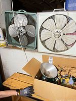Two box fans on ledge, cardboard box below containing light fixture, bulbs, string lights