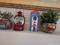 Four items on mantle: two small ceramic pots with faux plants, red lantern snow globe, unrelated shortbread tin