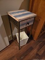 Full view of side table showing wood top, drawer, metal legs, and white plastic bin placed underneath