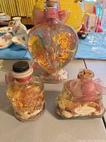 Front view of all three decorative bath oil bottles showing shapes, liquid color, botanicals, shells and closures