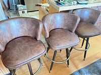 Front view of all three brown faux leather swivel bar stools showing seating surfaces, backs, legs, and footrest rings
