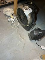 Three electrical items on basement floor: black air circulator fan, white clamp fan, black space heater