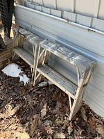 Two aluminum work stands leaning against siding with leaves and snow at base