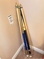 Four cue sticks leaning in corner; two BCE cues (black and blue wraps) are part of lot