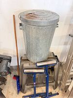 Galvanized trash can on plastic sawhorse, pair of blue roller stands visible, broom and dustpan nearby