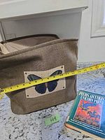 Front view of brown collapsible fabric bin with butterfly patch and stack of three paperback books, tape measure showing width ~14 in
