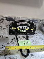 Front view of Alex's Bar metal sign with tape measure showing roughly 6 inch width
