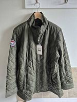 Front view of olive quilted jacket on hanger showing tags, zipper and snap placket, pockets