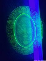 Full view of glowing green uranium glass platter under blacklight showing oval shape and floral etching