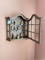 Cabinet on wall with door open showing multiple Hummel figurines on three shelves