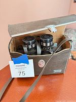 Binoculars stored in open brown case, view of eyepiece ends and neck strap