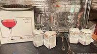 Full lot overview showing boxed Saint-Gobain glass set, loose stemware, ceramic spice canisters, metal utensils and plate