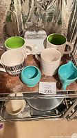 Six ceramic mugs arranged on shelf: two white ribbed green-interior, one white ribbed, one white script text, turquoise pedestal, turquoise spout mug