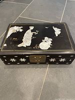 Full view of closed black lacquer jewellery box showing lid scene, floral inlay on front, metal clasp