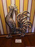 Full view of cast metal rooster wall decor against striped wall with ruler for scale