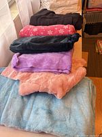Stack of colored plush throws in peach, light blue, purple, black, pink snowflake plus white throws behind