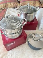 Two stainless steel Dutch ovens with lids on Parini boxes and gray nonstick saucepan with lid