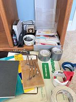 Wide view of assorted office supplies on floor including tape rolls, dispenser, columnar pad, letter holder, file holder, greeting card box