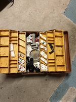 open tackle box showing wrapped lures, reel, bobbers, clamp tool, small container