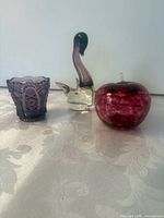Overall view of the three glass items: apple paperweight, swan figurine, purple votive holder