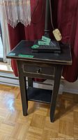 Front view of black distressed side table with lamp on top showing drawer, tapered legs, lower shelf