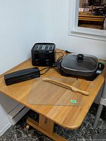 All items grouped on table: electric skillet with lid, 4-slice toaster, Hamilton Beach carving knife case, glass cutting board, wooden-handled bread knife