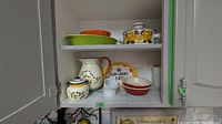 Cabinet shelf showing orange polka-dot bowl, green ribbed dish, yellow floral casserole, pitcher, sugar jar, ramekins, red bowl and weight plate