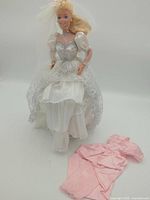 Full view Barbie doll seated with bridal dress and separate pink gown on side