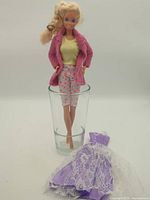 Full view of Barbie doll standing in glass with extra purple lace dress in front