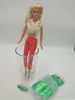 Full view of Barbie doll in glass with red pants, cream top, green accessories on surface