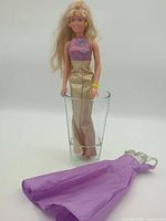 Full view of Maxie doll standing in glass wearing gold gown; purple dress laid beside