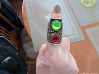 Ring worn on finger showing size, green and red cabochons, silver-plated finish