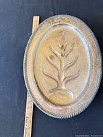 Front of oval brass plaque with tree relief and ruler for scale