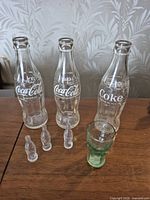 Three full-size clear Coca-Cola bottles, three clear mini bottle figurines, one green mini glass