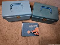 Two blue plastic storage boxes with handles and closed latches; Dennison Buttoneer box positioned in front