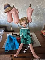 Doll seated on table with three mannequins, blue coat, straw hat, sunglasses, red hatbox visible