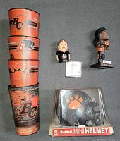 Group shot of all items: four cups stacked, Elimimian bobblehead, Passaglia figure with certificate, signed mini helmet in case