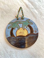 Front view of round glazed ceramic wall hanging showing palm tree and sunset design with cord