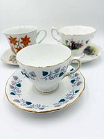 Three bone china teacup and saucer sets; front shows blue leaf pattern, rear shows orange lily and purple bouquet designs
