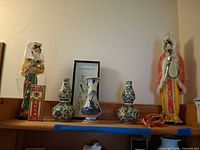 Overall lot on shelf showing two dolls, Delft vase, two gourd vases, framed art, decorative box