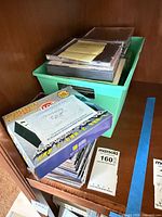 Shelf view showing green bin with stacked jewel cases, karaoke CD box set, loose CD stack